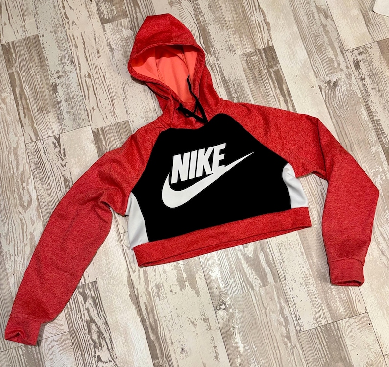 May include: Red and black cropped hoodie with the Nike logo in white. The hoodie has a hood and long sleeves. The body of the hoodie is black with the Nike logo, and the sleeves and hood are red.