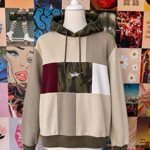 May include: A color-block hoodie with a camouflage hood and center panel. The hoodie features panels of beige, burgundy, and white, with the Nike logo embroidered on the camouflage section. The bottom hem and cuffs are olive green.