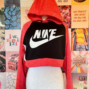 May include: Red and black cropped hoodie with the Nike logo in white. The hoodie has a drawstring hood and long sleeves. The background is a collage of colorful posters.
