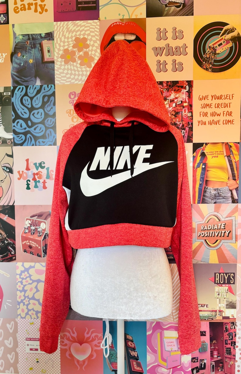 May include: Red and black cropped hoodie with the Nike logo in white. The hoodie has a drawstring hood and long sleeves. The background is a collage of colorful posters.