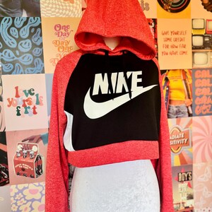 May include: A red and black Nike cropped hoodie with a white Nike logo. The hoodie has a hood and long sleeves. The background is a collage of colorful posters.