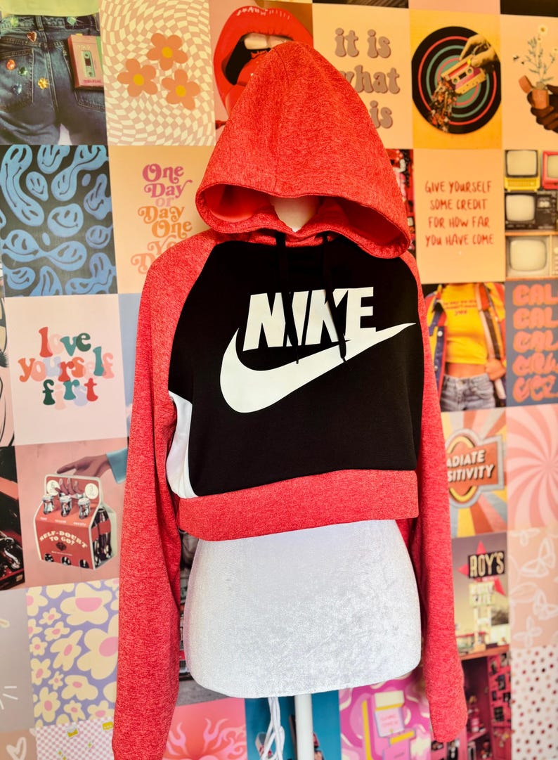 May include: A red and black Nike cropped hoodie with a white Nike logo. The hoodie has a hood and long sleeves. The background is a collage of colorful posters.