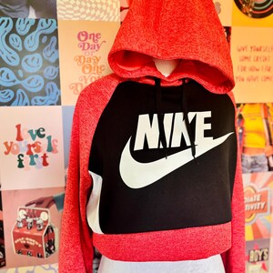May include: A red and black cropped hoodie with the Nike logo in white. The hoodie has a drawstring hood and long sleeves. The word "NIKE" is prominently displayed across the chest. The background features a collage of colorful posters.
