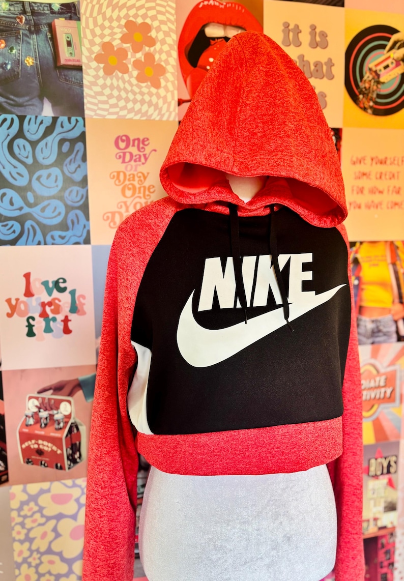 May include: A red and black cropped hoodie with the Nike logo in white. The hoodie has a drawstring hood and long sleeves. The word "NIKE" is prominently displayed across the chest. The background features a collage of colorful posters.