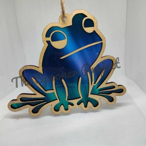 May include: A wooden frog ornament with a blue and green galaxy design. The frog is sitting on its hind legs with its front legs outstretched.