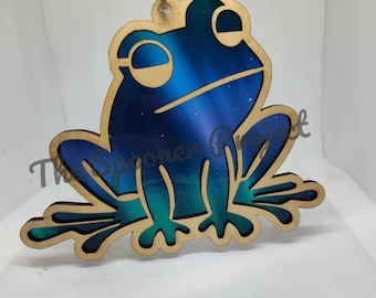 May include: A wooden frog ornament with a blue and green galaxy design. The frog is sitting on its hind legs with its front legs outstretched.