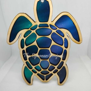 May include: A wooden sea turtle ornament with a blue, green, and purple ombre design. The turtle's shell is detailed with a geometric pattern. The ornament has a string for hanging.