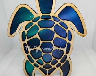 May include: A wooden sea turtle ornament with a blue, green, and purple ombre design. The turtle's shell is detailed with a geometric pattern. The ornament has a string for hanging.