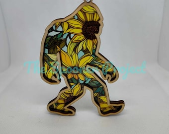 May include: A wooden ornament shaped like a Bigfoot, covered in a sunflower pattern. The ornament is painted in shades of yellow, green, and brown.