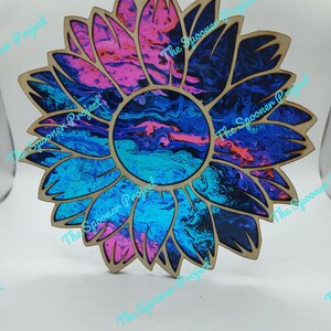 Könnte beinhalten: Holz-Sonnenblumen-Ornament mit einem wirbelnden Muster in Blau-, Rosa- und Lilatönen. Die Sonnenblume hat einen natürlichen Holzrand und ein Loch zum Aufhängen. Der Text "The Spooner Project" ist sichtbar.