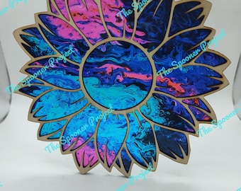 May include: Wooden sunflower ornament with a vibrant, swirling pattern in shades of blue, pink, and purple. The sunflower has a natural wood border and a hole for hanging. The text "The Spooner Project" is visible.