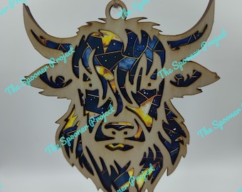 May include: A wooden ornament shaped like a Highland cow's head with a blue, black, and gold galaxy-like pattern. The ornament has a string for hanging.