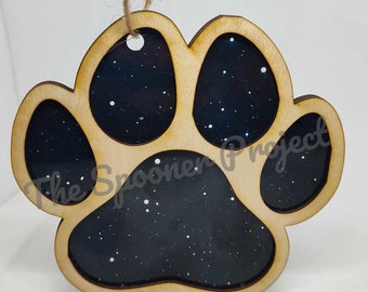 May include: Wooden paw print ornament with a black and white galaxy design. The ornament has a twine hanger and the text "The Spooner Project" is visible on the bottom.