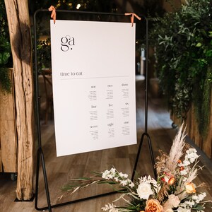 May include: A black metal framed sign with a white seating chart, featuring the text "ga" and "time to eat." The sign is adorned with orange ribbons and rests on a wooden floor. A floral arrangement with white, peach, and beige flowers is at the base.