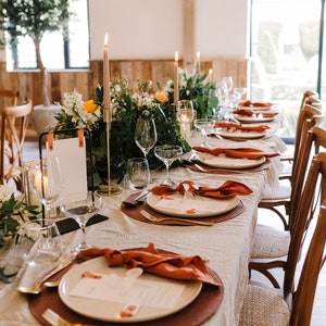 May include: Elegant table setting for a special event. The table is covered with a cream-colored linen tablecloth and set with white plates, rust-colored napkins, and gold cutlery. Tall candles and floral arrangements add to the ambiance.
