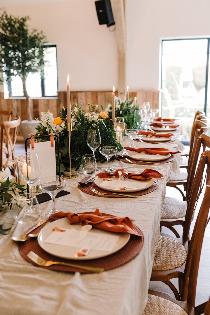 May include: Elegant table setting for a special event. The table is covered with a cream-colored linen tablecloth and set with white plates, rust-colored napkins, and gold cutlery. Tall candles and floral arrangements add to the ambiance.