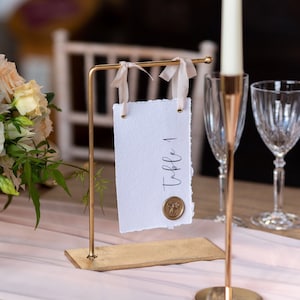 Könnte beinhalten: Ein goldfarbener Tischkartenhalter mit einer weißen Karte mit der Aufschrift "Table 1". Die Karte ist mit einem Wachssiegel versehen und mit Bändern gebunden. Ein Blumenstrauß und ein Kerzenhalter befinden sich im Hintergrund.