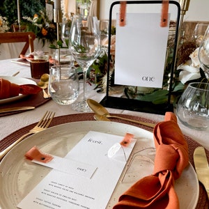 May include: Elegant table setting with a black metal sign displaying the word "one" and a menu card. The table is set with gold cutlery, white plates, and rust-colored napkins and ribbons. The menu card reads "feast."