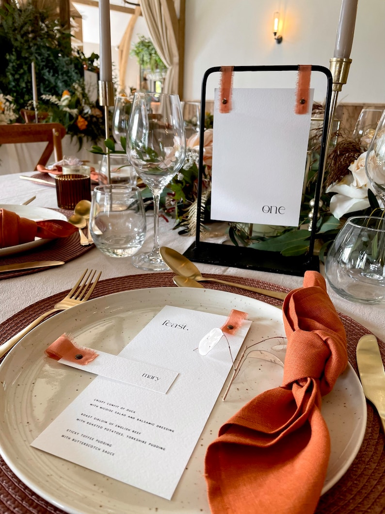 May include: Elegant table setting with a black metal sign displaying the word "one" and a menu card. The table is set with gold cutlery, white plates, and rust-colored napkins and ribbons. The menu card reads "feast."