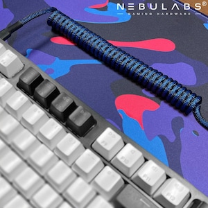 Könnte beinhalten: Eine weiße Tastatur mit schwarzen Tasten und einem blauen Spiralkabel auf einer bunten Mausmatte mit dem Text "NEBULABS® GAMING HARDWARE".