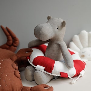 May include: A gray fabric hippopotamus wearing a red and white life preserver sits on a white surface next to a brown fabric crab.