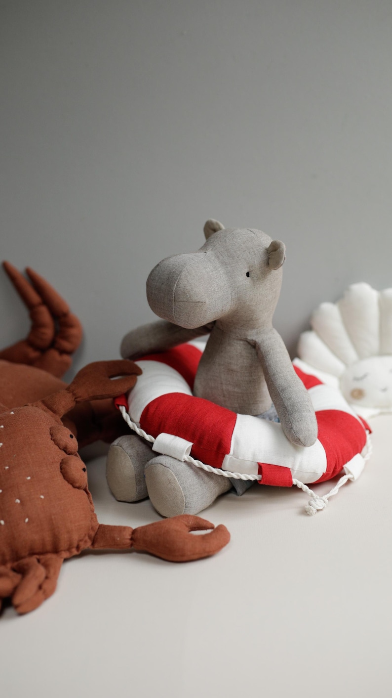 May include: A gray fabric hippopotamus wearing a red and white life preserver sits on a white surface next to a brown fabric crab.