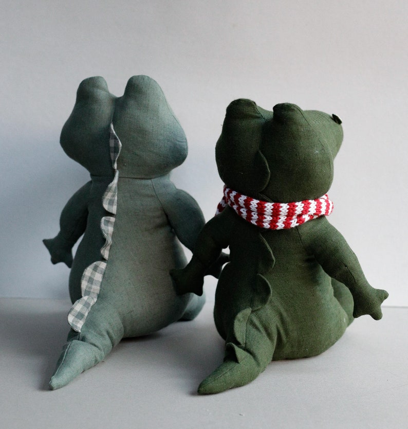May include: Two green fabric alligator plushies. One alligator is wearing a red and white striped scarf.