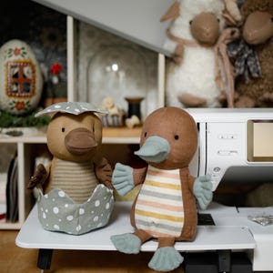May include: Two brown and beige plush platypus toys. One platypus is sitting in a gray and white polka dot egg. The other platypus is sitting on a white surface with its feet outstretched.