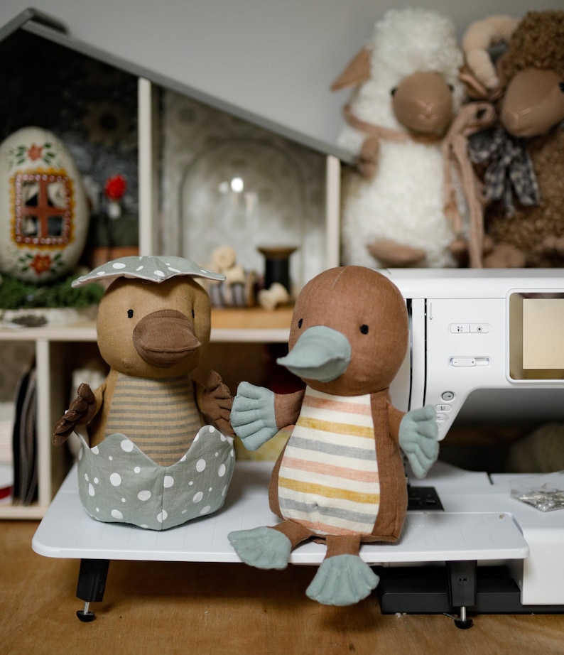 May include: Two brown and beige plush platypus toys. One platypus is sitting in a gray and white polka dot egg. The other platypus is sitting on a white surface with its feet outstretched.