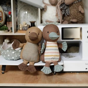 May include: Two brown and beige striped plush platypus toys with blue beaks and feet. The toys are sitting on a white surface in front of a white sewing machine.