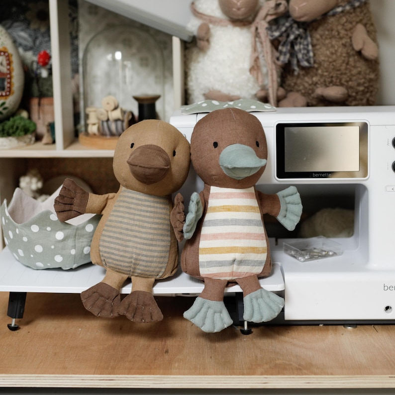 May include: Two brown and beige striped plush platypus toys with blue beaks and feet. The toys are sitting on a white surface in front of a white sewing machine.