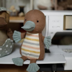 May include: A brown and blue plush toy shaped like a platypus. The toy has a striped body and a long, flat beak.