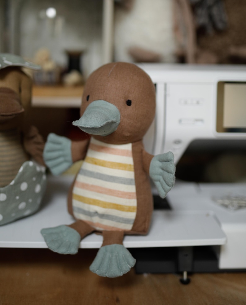 May include: A brown and blue plush toy shaped like a platypus. The toy has a striped body and a long, flat beak.