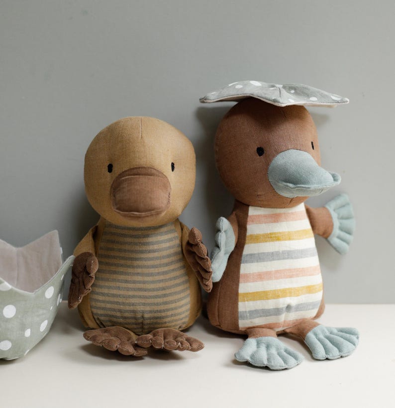 May include: Two plush toy duck-billed platypuses. One is brown with a gray and yellow striped shirt and a gray hat. The other is brown with a gray and pink striped shirt. Both have blue feet.