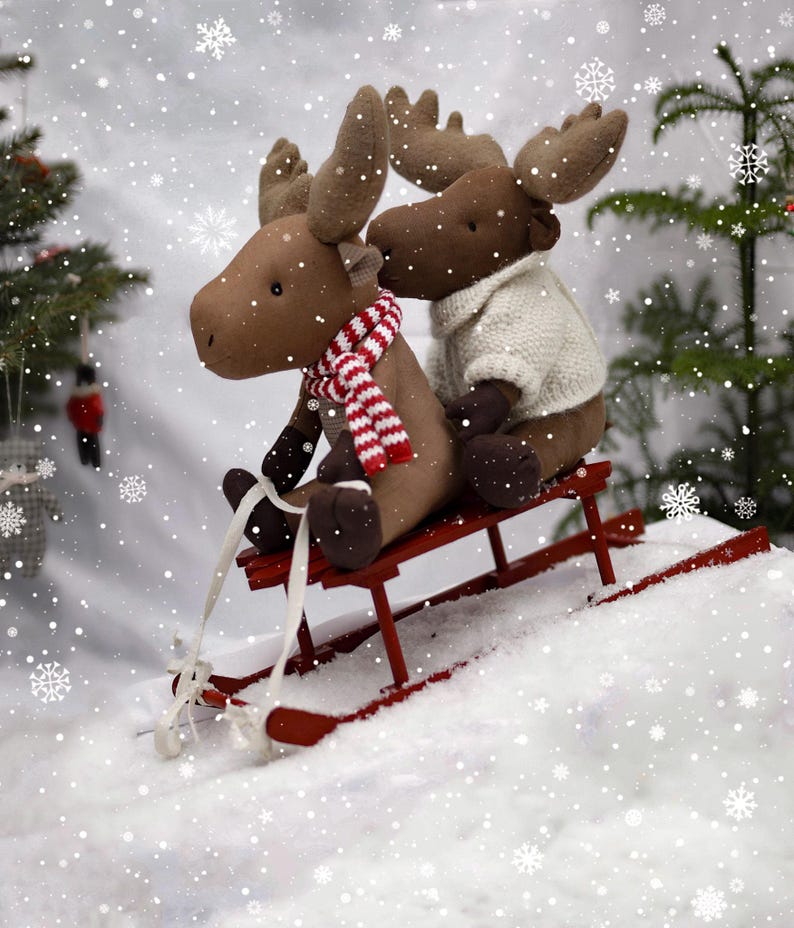 May include: Two brown plush moose toys riding on a red wooden sled in a snowy winter scene.