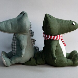 May include: Two plush crocodile toys, one green and one green with a red and white striped scarf. The crocodiles are sitting side by side.