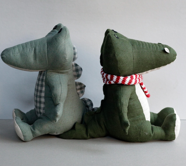 May include: Two plush crocodile toys, one green and one green with a red and white striped scarf. The crocodiles are sitting side by side.