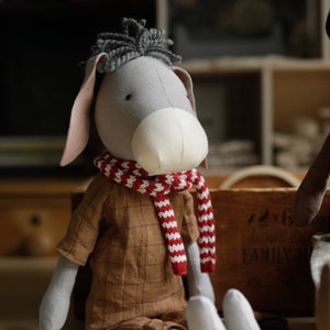 Puede incluir: Un burro de peluche gris que lleva una bufanda a rayas rojas y blancas y una camisa de cuadros marrones. El burro est&aacute; sentado sobre una superficie de madera.