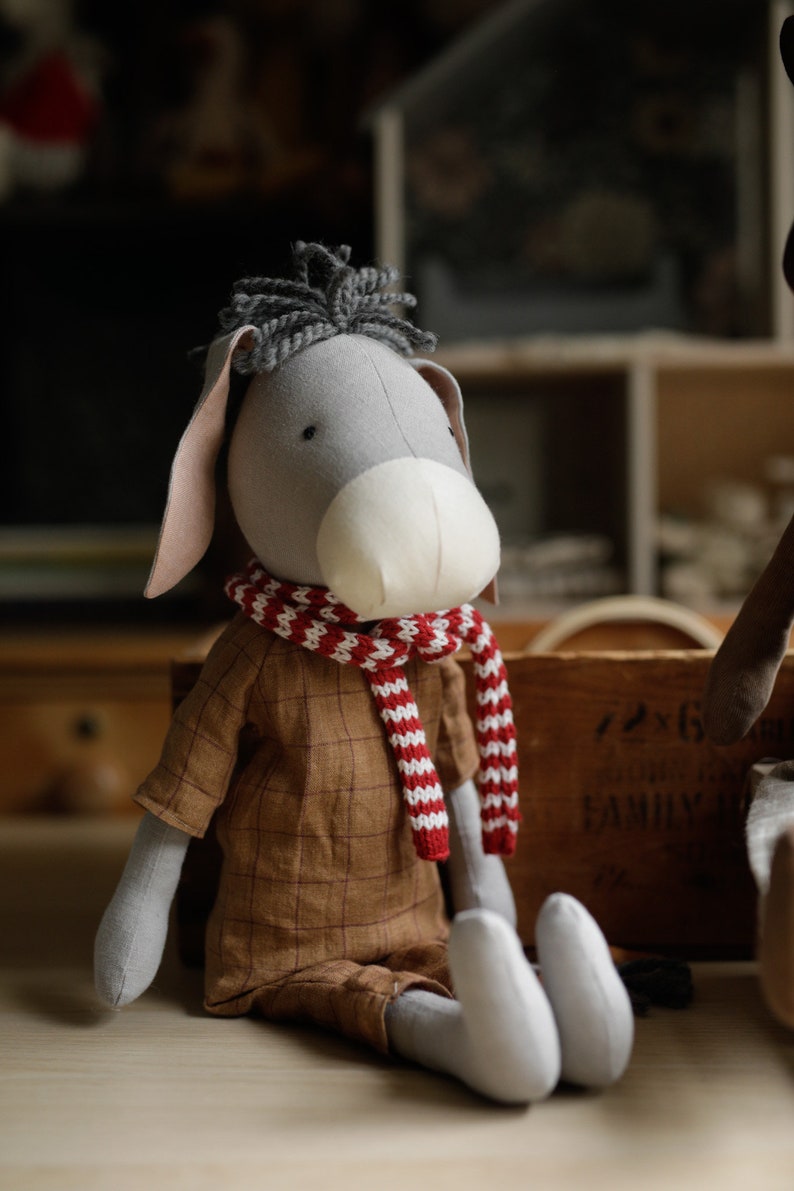 Puede incluir: Un burro de peluche gris que lleva una bufanda a rayas rojas y blancas y una camisa de cuadros marrones. El burro est&aacute; sentado sobre una superficie de madera.