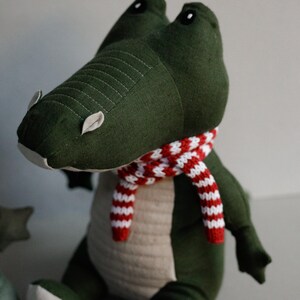 May include: Green plush crocodile toy wearing a red and white striped scarf. The crocodile has black eyes and a stitched mouth.