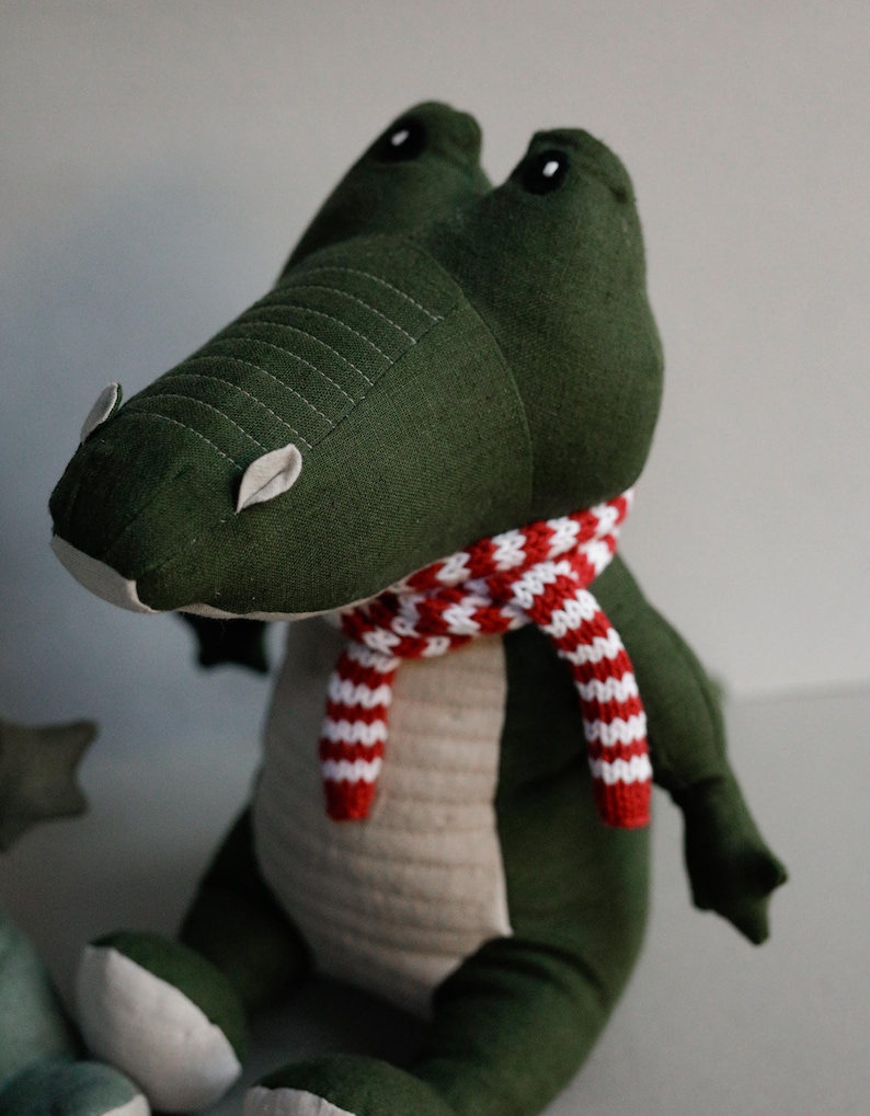 May include: Green plush crocodile toy wearing a red and white striped scarf. The crocodile has black eyes and a stitched mouth.