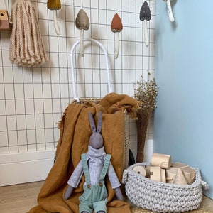 Puede incluir: Un conejo de peluche gris con un mono est&aacute; sentado en una manta marr&oacute;n. Una cesta de mimbre blanca con asa est&aacute; detr&aacute;s del conejo. Una guirnalda de setas de fieltro cuelga de la pared detr&aacute;s de la cesta. Una cesta de ganchillo con bloques de madera est&aacute; en primer plano.
