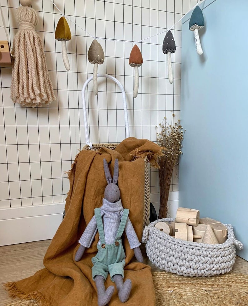 Puede incluir: Un conejo de peluche gris con un mono est&aacute; sentado en una manta marr&oacute;n. Una cesta de mimbre blanca con asa est&aacute; detr&aacute;s del conejo. Una guirnalda de setas de fieltro cuelga de la pared detr&aacute;s de la cesta. Una cesta de ganchillo con bloques de madera est&aacute; en primer plano.