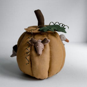 May include: A stuffed fabric pumpkin in a muted mustard yellow color featuring a dark brown stem and a green leaf. Attached to the pumpkin are small plush animal heads including a deer with white spots, a hedgehog, and a duck, with a curly vine.