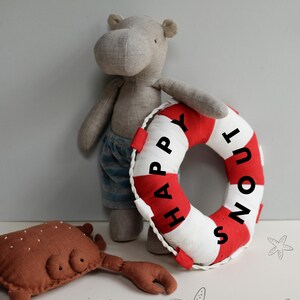 May include: A gray stuffed hippopotamus holding a red and white life preserver with the text "Happy Snout" on it. A brown stuffed crab is in the foreground.