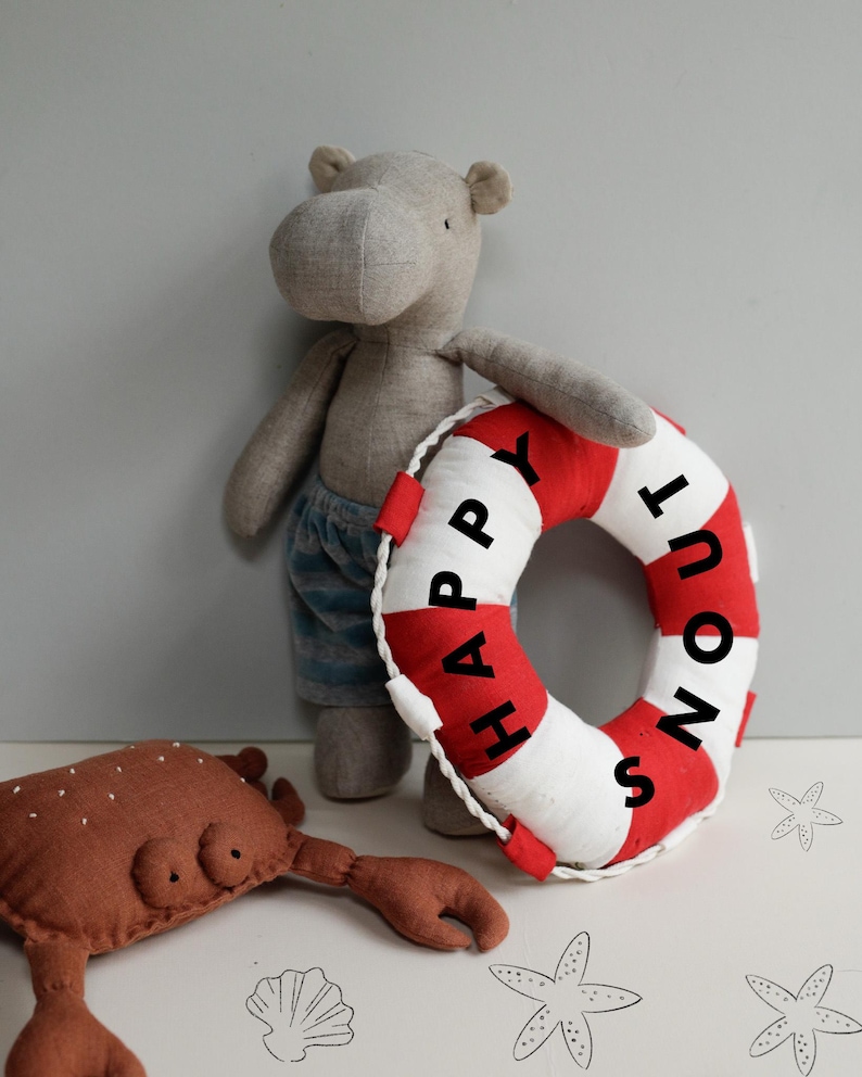 May include: A gray stuffed hippopotamus holding a red and white life preserver with the text "Happy Snout" on it. A brown stuffed crab is in the foreground.