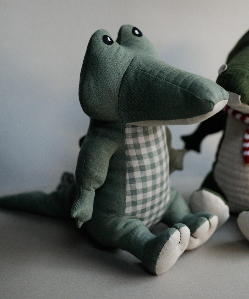 May include: A green plush crocodile toy with a white and green checkered pattern on its chest. The crocodile is sitting with its legs crossed and has a long tail.