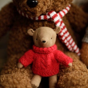 May include: A brown teddy bear wearing a red and white striped scarf is holding a smaller brown teddy bear wearing a red knitted sweater.