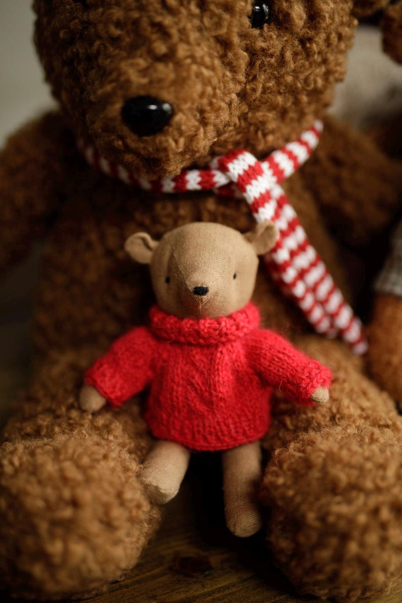 May include: A brown teddy bear wearing a red and white striped scarf is holding a smaller brown teddy bear wearing a red knitted sweater.