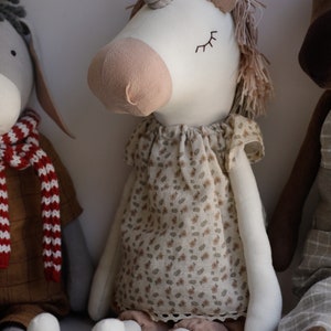 Puede incluir: Un juguete de peluche de unicornio blanco con la nariz rosa y los ojos cerrados. El unicornio lleva un vestido floral marr&oacute;n y blanco y pantalones marrones. El unicornio est&aacute; sentado en una superficie gris.